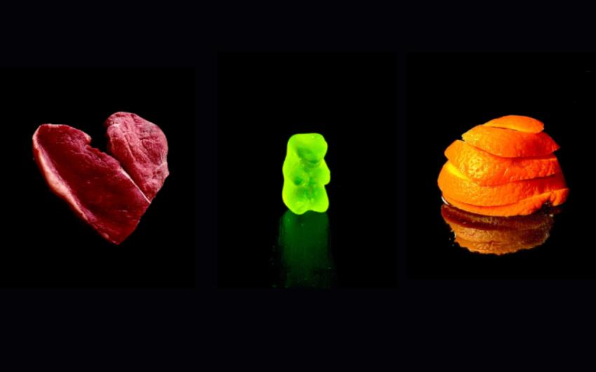 Black void with images of a raw steak in the shape of a heart, a yellow gummybear, and an orange peel arranged in a spiral.