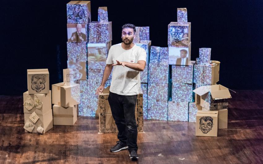 A mixed race man in his 30s, wearing black jeans and a white T-Shirt, crosses the stage in front of a wall of brown cardboard boxes, onto which is projected images of politicians and TV static