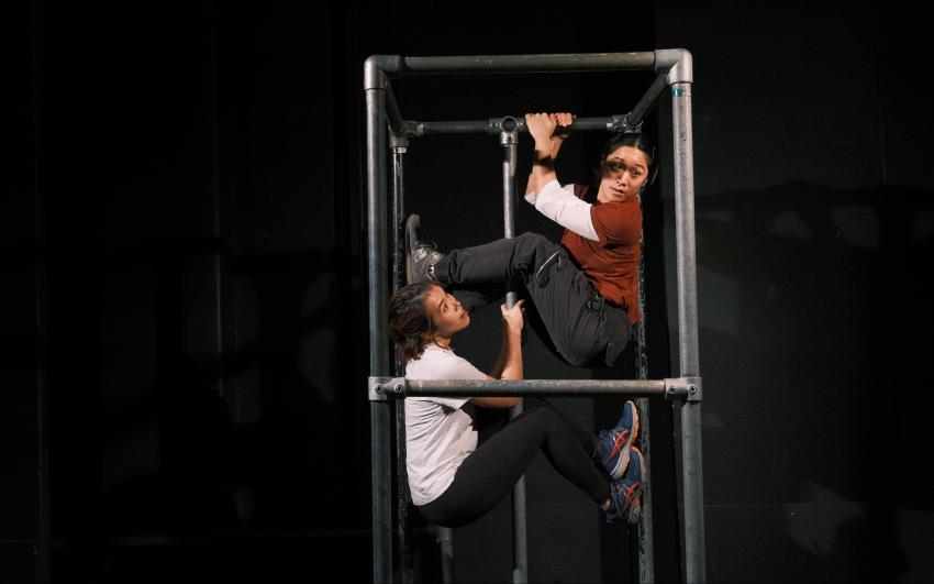  Isabella (East Asian woman) and Louiseanne (East Asian nonbinary person) are wedged inside a silver scaffolding frame on stage, looking out to the audience.