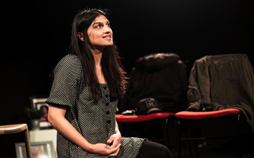 A woman sits on the floor about to speak. She surrounded by laundry. A bright stage spotlight shines above her.