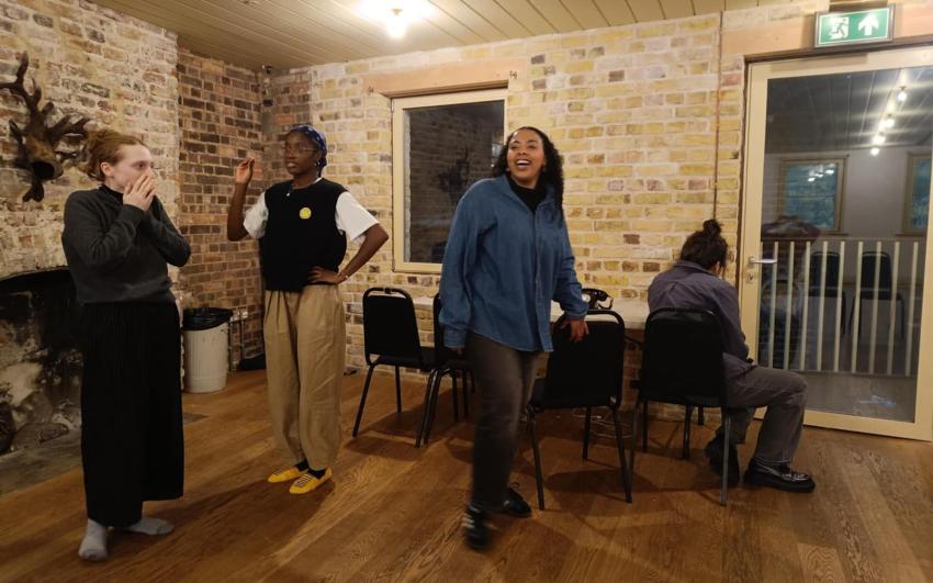 Four young women are in a brick walled room with a wooden floor. One woman has her hands to her face, another has an arm raised with a hand on her hip. The woman in the centre of the picture s standing and smiling. One woman is sat facing way from the cam