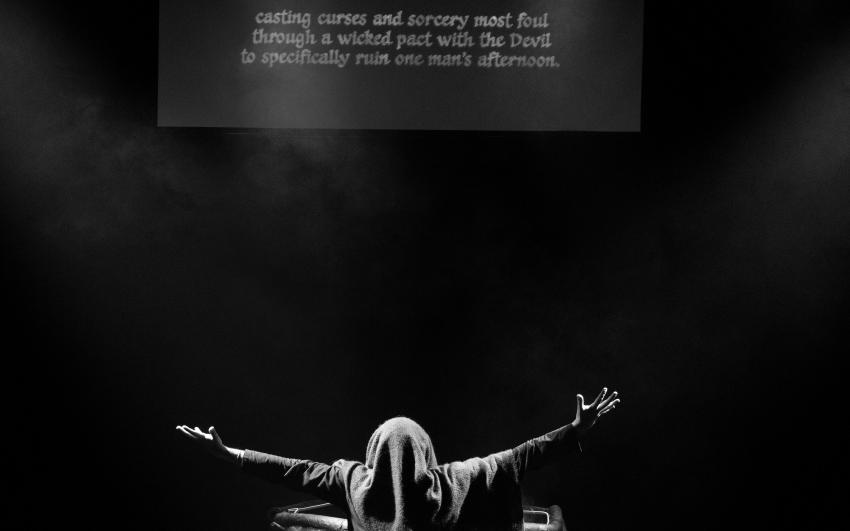 Edy Hurst, a white man in his early 30s with thick rimmed glasses holds his arms out on stage as captions appear on a projector above him