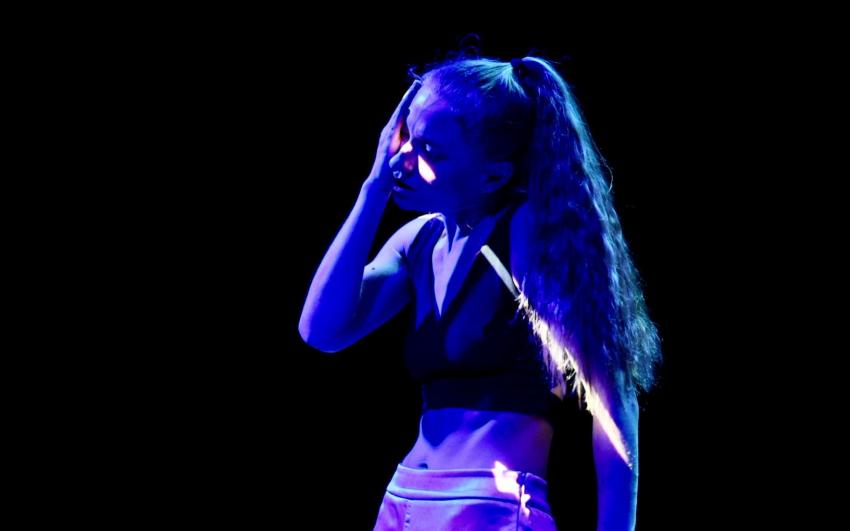 A woman with blonde hair and a black top stands in blue light with her right hand placed on her cheek suggesting a moment of reflection or physical exertion