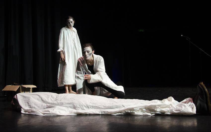 Two people dressed in white standing over a third person who is draped in a white sheet. 