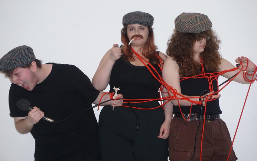 Three people wearing flatcaps all tangled in red rope. One person is looking at a plastic toy cow, another is lighting a cigarette, and another is looking at the rope tangled on their arm