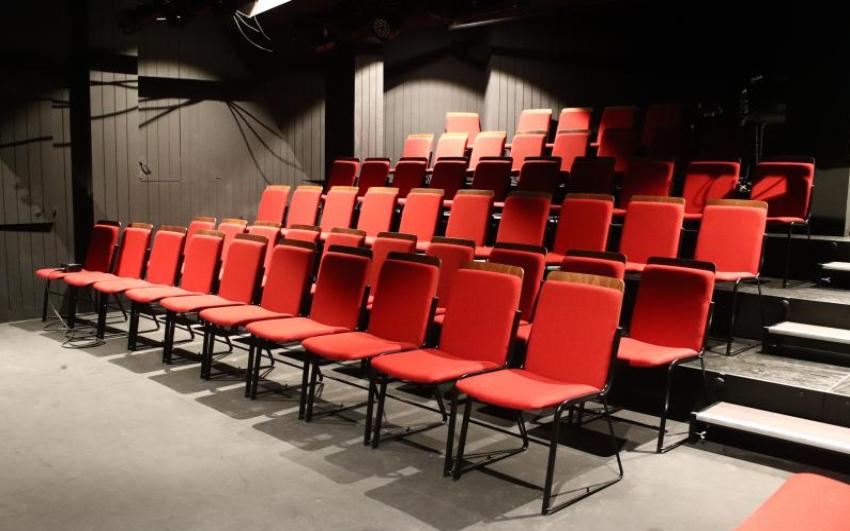 Rows of chairs with red fabric seats, arranged in rows on a raked seating bank, sitting within a black box theatre. 