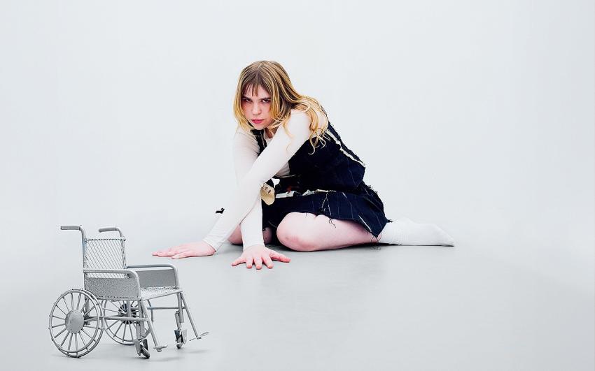 A young woman wearing childlike clothing sat on the floor next to an empty wheelchair.