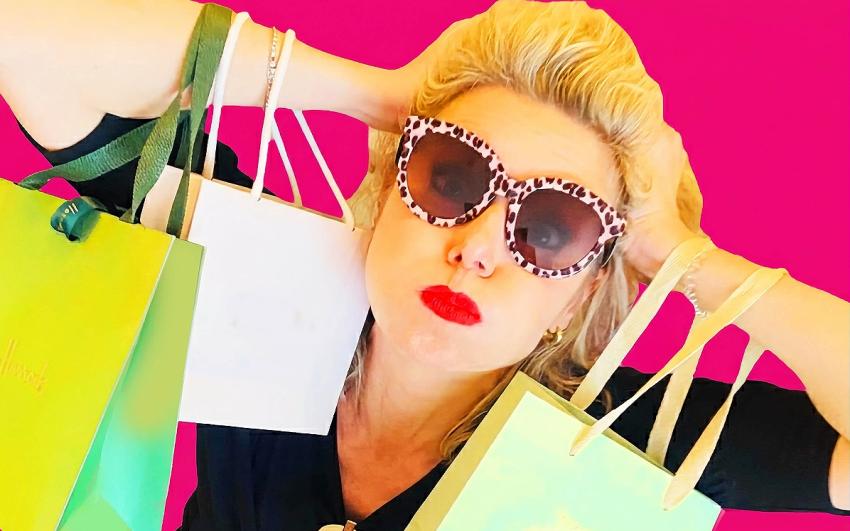 Woman wearing leopard-print sunglasses and red lipstick poses against a bright pink background, holding multiple shopping bags, swamped by consumer goods—playful yet overwhelmed.