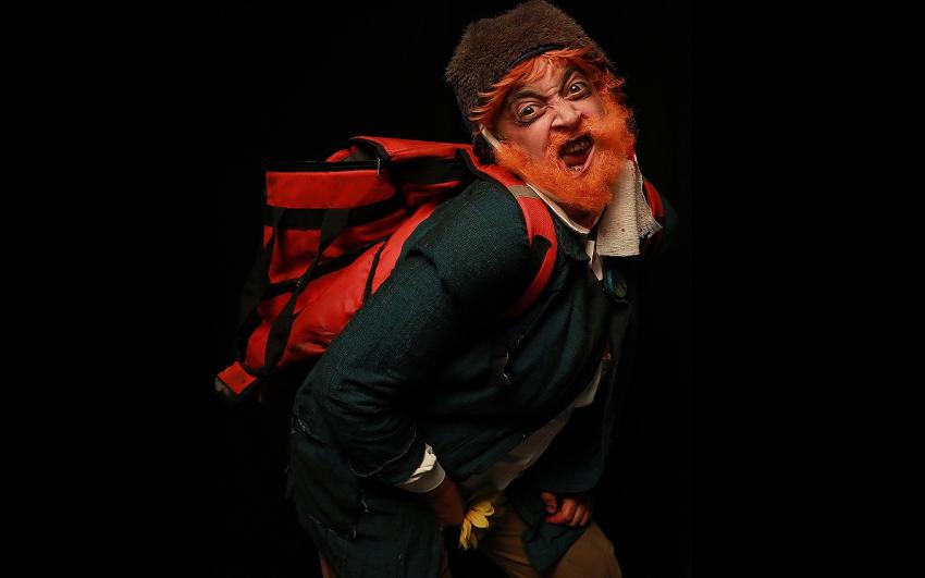 An man with orange hair, a scraggly beard, a bandage around his head and a food delivery backpack makes a distorted face toward the camera.