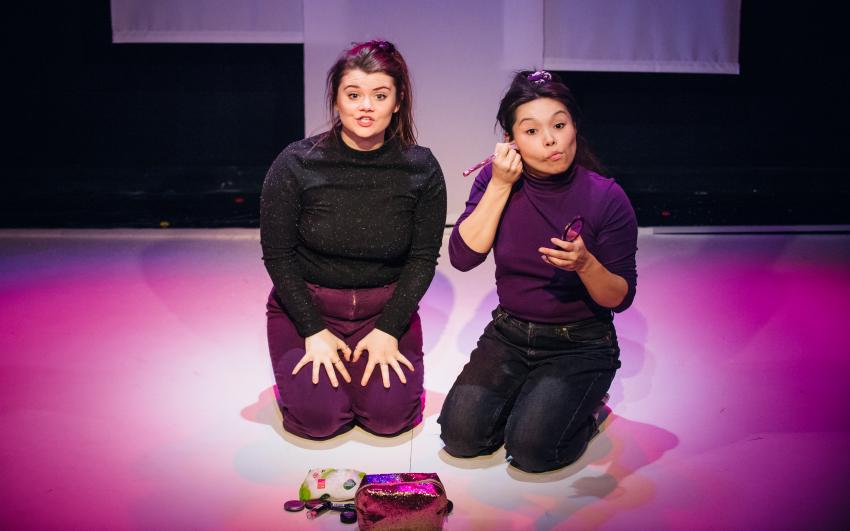 A photo of two actresses, kneeling on a purple stage. One is talking and the other is doing makeup