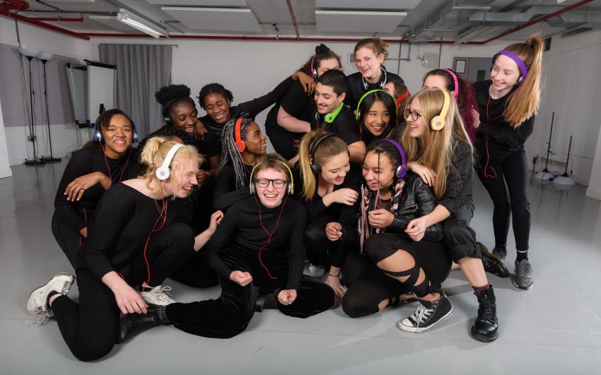A photo of teenagers in black clothes, laughing in a huddle, they are all wearing colour headphones