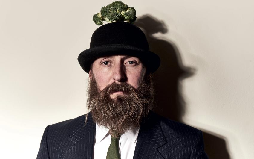 A bearded man with a suit and bowler hat faces the camera, there is broccoli on his head