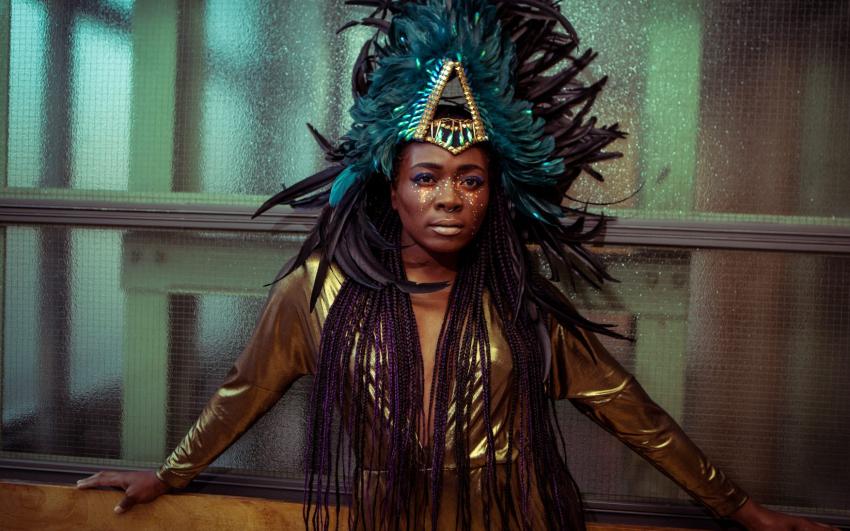 A young woman looks towards the camera she is wearing a dark green headress on with gold makeup 